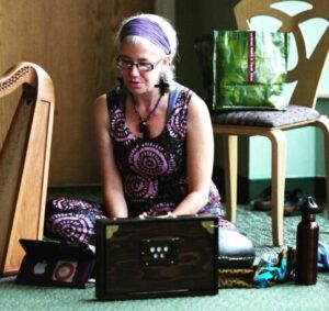 Image of Rachel, a white woman with gray hair playing a shruti box. A Celtic harp sits on the floor beside her.