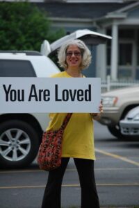 Rachel, a middle aged woman of medium build, wearing yellow tshirt and holding a rectangle large sign that says YOU ARE LOVED in block letters